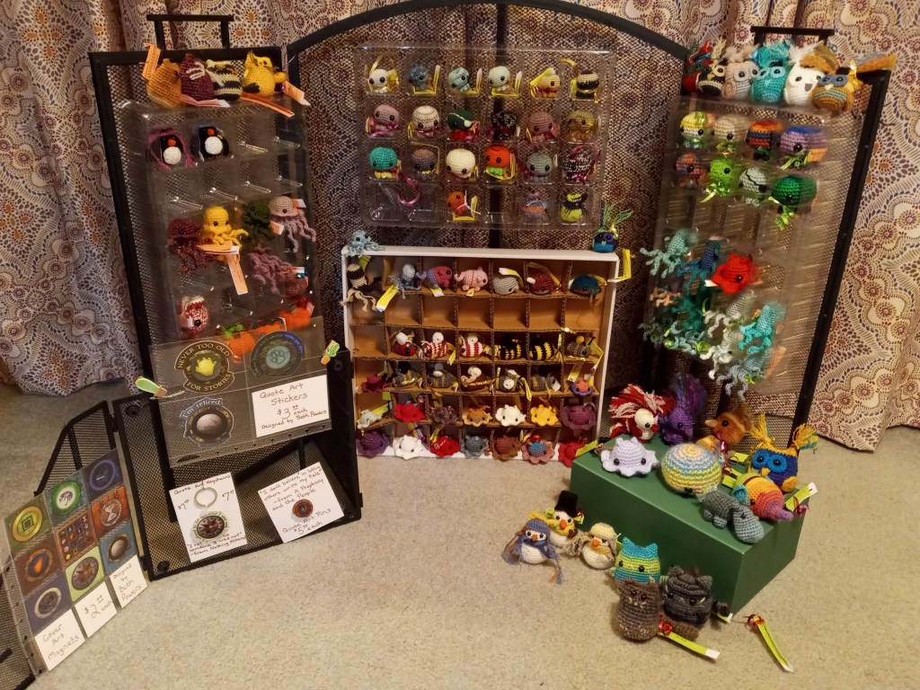 Large trifold wire display with individual compartments housing small crochet critters.A smaller wire display holding magnets, stickers, a keychain, and pin along with a box supporting larger crochet critters in front. 